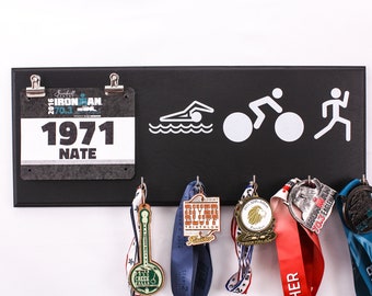 May include: Black wooden medal display rack with hooks for hanging medals. The rack has a white silhouette of a swimmer, cyclist, and runner. A black and white race bib with the number 1971 and the name Nate is clipped to the top of the rack. There are six medals hanging from the hooks.