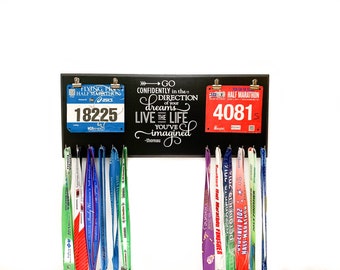 May include: Black medal display rack with hooks for hanging medals and two race bibs. The bibs are blue and red with white numbers. The rack has a quote that reads "Go confidently in the direction of your dreams. Live the life you've imagined - Thoreau".