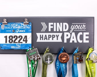 May include: A gray wooden medal display with the text "FIND your HAPPY PACE" in white. The display has hooks for hanging medals and a clip for holding a race bib. A blue race bib with the number "18224" is clipped to the display. Several medals with colorful ribbons are hanging from the hooks.