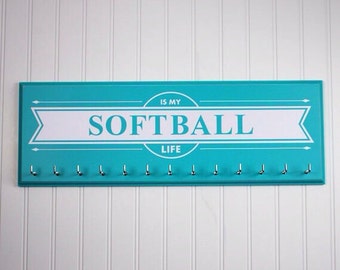 May include: A turquoise blue wooden sign with the text "Softball is my life" in white lettering. The sign has 12 silver hooks for hanging medals or other items.