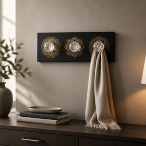 May include: A black wall-mounted coat rack with three crystal knobs and gold mandala designs. A beige scarf hangs from one of the knobs. The rack is mounted on a neutral-colored wall, with a dark brown dresser in the foreground.