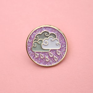 May include: A purple glitter enamel pin featuring a white and grey cloud with raindrops falling from it.