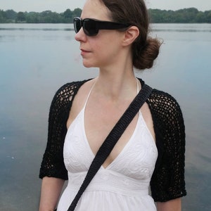 May include: A black crochet shrug with short sleeves and a matching black shoulder bag. The woman is wearing a white dress and sunglasses. The image is taken outdoors near a body of water.