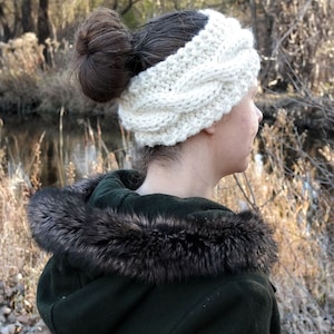 May include: A cream-colored, cable-knit headband. The thick, textured ear warmer is worn on the head. The person is wearing a dark green coat with a fur-trimmed hood. The background features a natural outdoor setting with trees and a body of water.