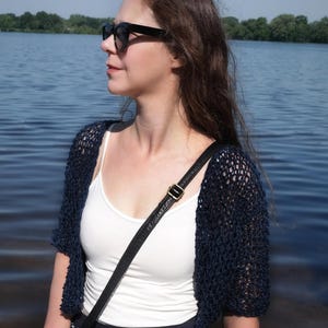 May include: A dark navy blue open-knit crochet bolero cardigan with short sleeves, worn over a white camisole and a navy blue skirt with white polka dots. A black crossbody bag with a gold-tone buckle strap is worn over the shoulder.