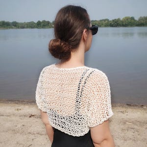May include: A cream-colored, open-weave crochet top with short sleeves. The top is worn over a black garment. The word "ORNUTE" is visible at the bottom of the image. The background features a lake and trees.