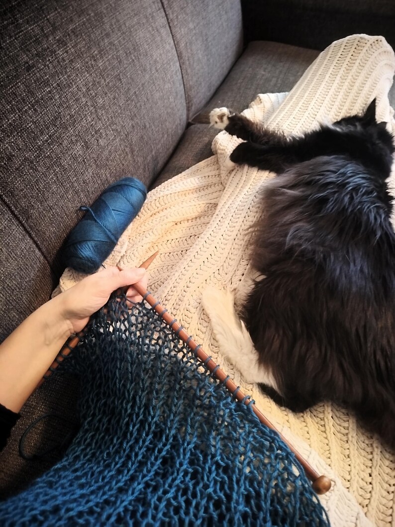 May include: A person knitting with blue yarn and wooden needles. A ball of blue yarn sits nearby. A black cat rests on a cream-colored knitted blanket on a gray sofa. The knitted project is a gradient of blue.