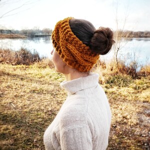 Pu&ograve; includere: Una donna indossa una fascia per capelli lavorata a maglia color giallo senape e un maglione a collo alto color crema. La fascia &egrave; spessa e strutturata, e il maglione ha un motivo a coste. Lo sfondo mostra un lago e alberi.