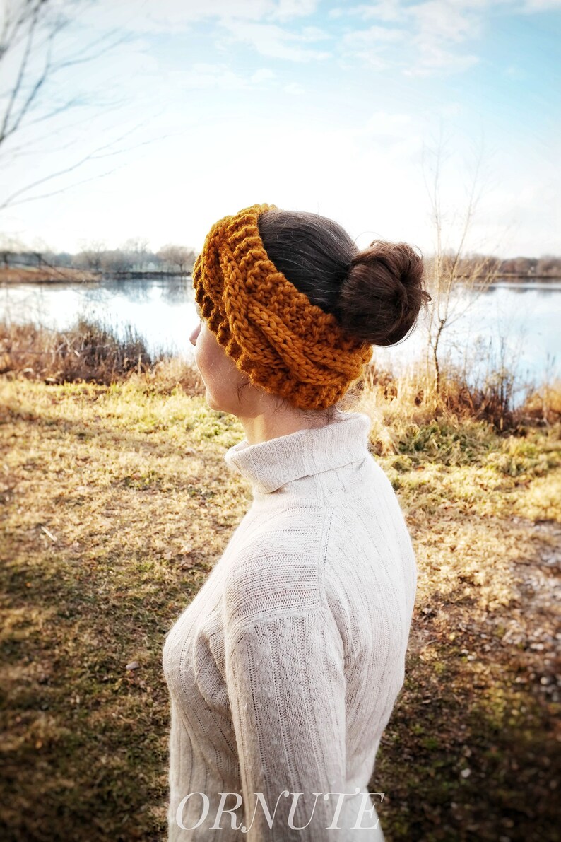 Pu&ograve; includere: Una donna indossa una fascia per capelli lavorata a maglia color giallo senape e un maglione a collo alto color crema. La fascia &egrave; spessa e strutturata, e il maglione ha un motivo a coste. Lo sfondo mostra un lago e alberi.