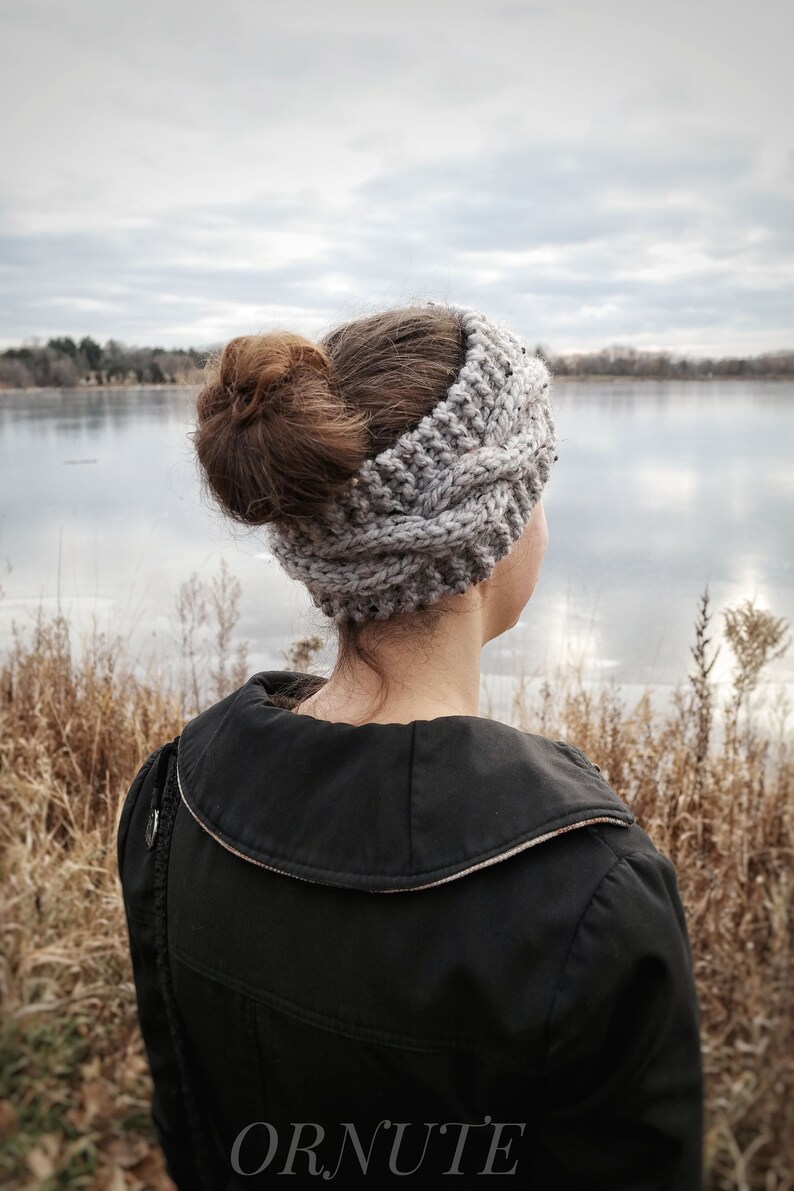 Pu&ograve; includere: Una fascia grigia lavorata a maglia con un motivo a trecce. La fascia &egrave; indossata da una persona con i capelli raccolti in uno chignon. La persona indossa una giacca nera e guarda un lago. La parola "ORNUTE" &egrave; stampata in basso.