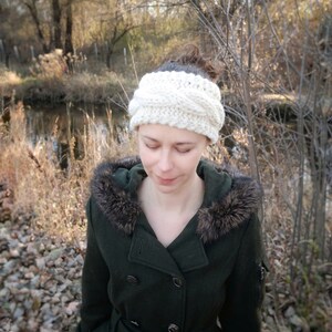 Pu&ograve; includere: Una fascia per capelli a trecce color crema. La fascia &egrave; spessa e sembra essere lavorata a mano. La persona indossa un cappotto verde scuro con cappuccio bordato di pelliccia. La parola "ORNUTE" &egrave; visibile in basso nell'immagine.