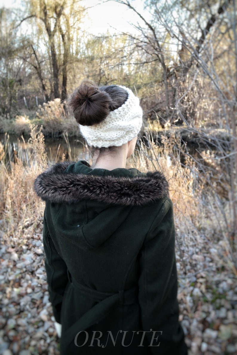 Pu&ograve; includere: Una donna indossa una fascia in maglia bianca con uno chignon marrone scuro. Indossa un cappotto verde scuro con cappuccio foderato in ecopelliccia. L'immagine &egrave; scattata all'aperto con uno sfondo naturale di alberi e un ruscello.
