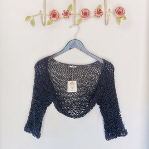 May include: A charcoal-colored, open-weave shrug hangs on a black hanger. The shrug has long sleeves and a loose, airy design. A decorative metal wall hook with pink roses and green leaves is above the shrug. The word "CHARCOAL" is at the bottom.