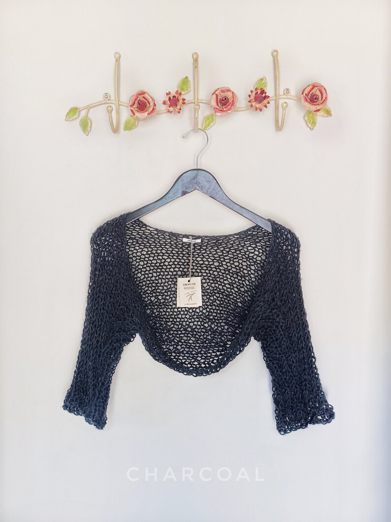 May include: A charcoal-colored, open-weave shrug hangs on a black hanger. The shrug has long sleeves and a loose, airy design. A decorative metal wall hook with pink roses and green leaves is above the shrug. The word "CHARCOAL" is at the bottom.