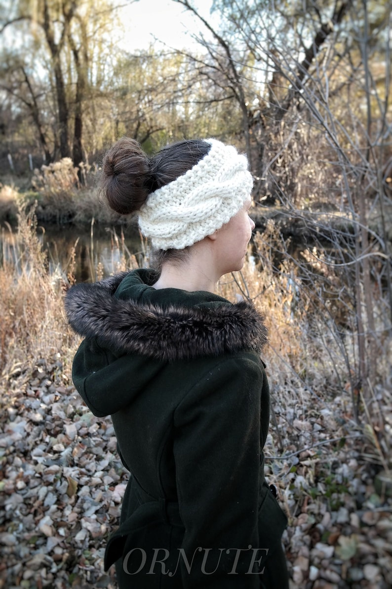 May include: A cream-colored, cable-knit headband. The thick, textured ear warmer is worn on the head. The person is wearing a dark green coat with a fur-trimmed hood. The background features a natural outdoor setting with trees and a body of water.