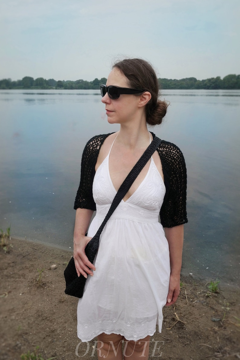 May include: A black crochet shrug with short sleeves and a matching black shoulder bag. The woman is wearing a white dress and sunglasses. The image is taken outdoors near a body of water.