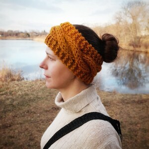 Pu&ograve; includere: Fascia per capelli lavorata a maglia color giallo senape. La fascia &egrave; realizzata con filato spesso e presenta un design a coste strutturato. La persona indossa un dolcevita color crema e ha i capelli raccolti in uno chignon. Lo sfondo comprende un lago e alberi.
