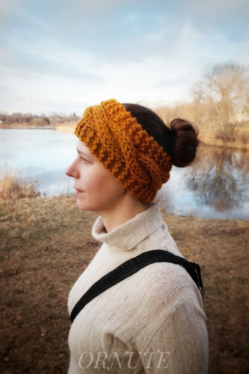 Pu&ograve; includere: Fascia per capelli lavorata a maglia color giallo senape. La fascia &egrave; realizzata con filato spesso e presenta un design a coste strutturato. La persona indossa un dolcevita color crema e ha i capelli raccolti in uno chignon. Lo sfondo comprende un lago e alberi.
