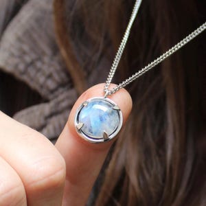 May include: A silver necklace with a round, light blue gemstone pendant. The pendant is set in a silver bezel and hangs from a delicate chain. The gemstone has a cloudy, ethereal appearance.