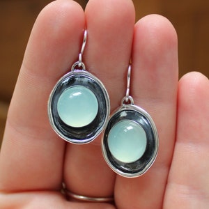 May include: A pair of silver earrings with a light blue gemstone set in a silver bezel. The earrings have a simple, elegant design.
