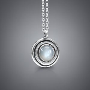 May include: A silver pendant necklace featuring a pale, round gemstone set in a textured, circular frame. The pendant is suspended from a fine silver chain, offering a simple, elegant design. The necklace is shown against a grey gradient background.