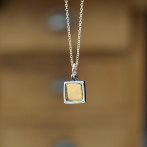 May include: A silver chain necklace featuring a small, square-shaped pendant. The pendant has a textured, gold-colored center framed by a smooth, polished silver border. The piece hangs vertically from a delicate, thin silver link chain.