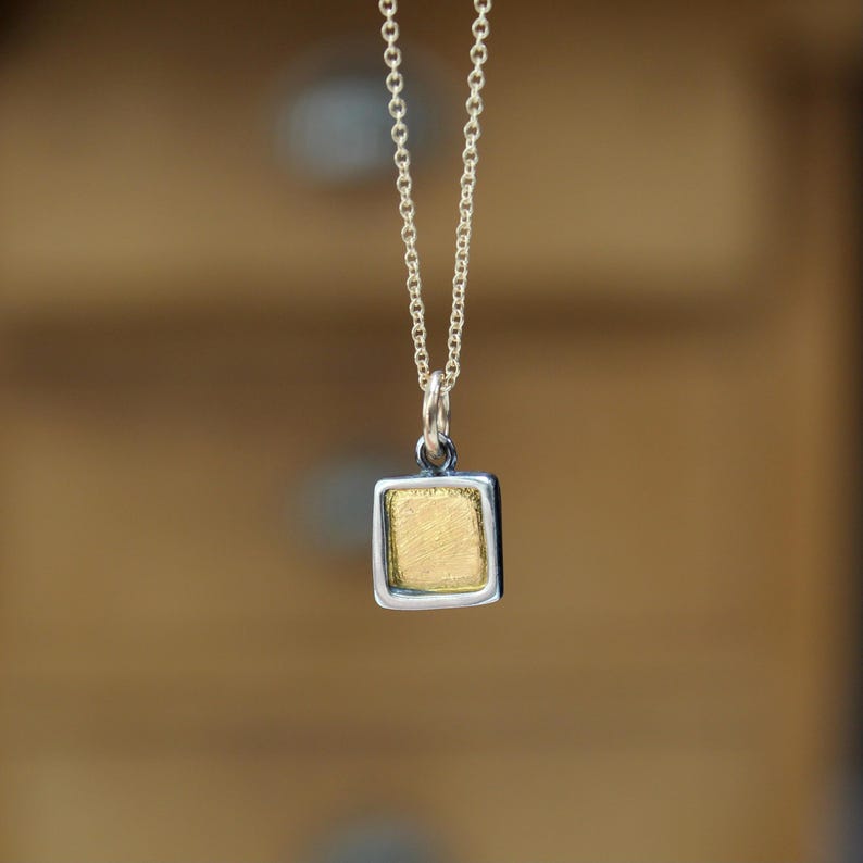 May include: A silver chain necklace featuring a small, square-shaped pendant. The pendant has a textured, gold-colored center framed by a smooth, polished silver border. The piece hangs vertically from a delicate, thin silver link chain.