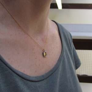May include: A delicate gold chain necklace featuring a small, rectangular pendant with a dark metallic frame and a bright gold-colored center. The pendant hangs from a simple jump ring attached to the thin, polished gold-tone metal chain.