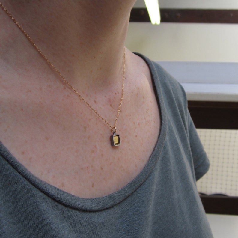 May include: A delicate gold chain necklace featuring a small, rectangular pendant with a dark metallic frame and a bright gold-colored center. The pendant hangs from a simple jump ring attached to the thin, polished gold-tone metal chain.