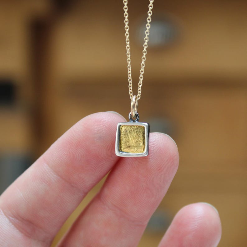 May include: A small square pendant necklace featuring a textured gold-colored center framed by a smooth silver-colored border. The pendant hangs from a delicate gold-colored chain, creating a simple and refined piece of jewelry for everyday wear.