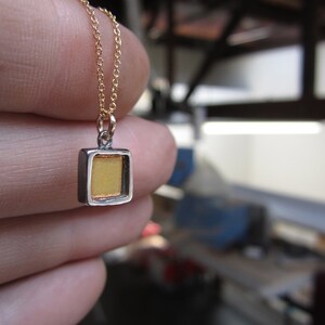 May include: A small square pendant necklace featuring a metallic silver frame with a recessed, textured gold-colored center. The pendant hangs from a delicate, thin gold-toned chain, showcasing a contrast between the silver edges and the gold inlay.