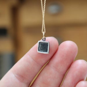 May include: A small square pendant with a dark, recessed center and a polished silver-toned border, suspended from a delicate gold-toned chain. The pendant features a simple geometric shape and a smooth, metallic finish on the outer frame.