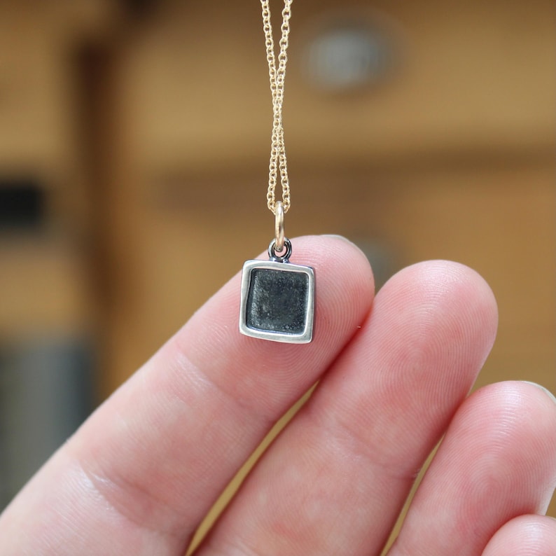 May include: A small square pendant with a dark, recessed center and a polished silver-toned border, suspended from a delicate gold-toned chain. The pendant features a simple geometric shape and a smooth, metallic finish on the outer frame.