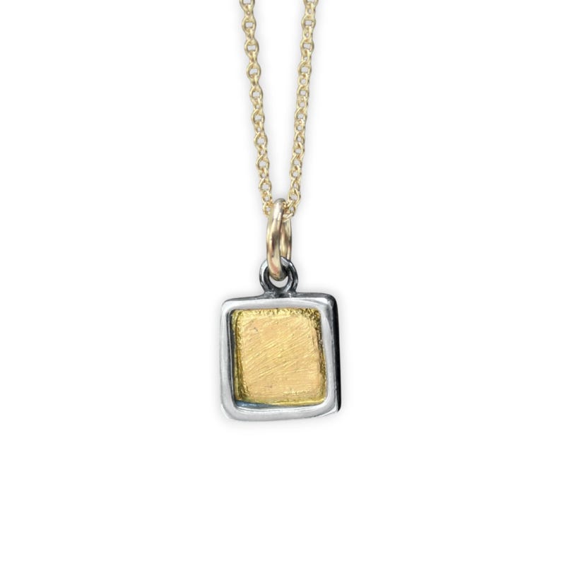 May include: A necklace featuring a square pendant with a textured gold-colored center framed by a smooth silver-colored border. The pendant hangs from a delicate gold-colored chain that connects to a small jump ring at the top of the square frame.
