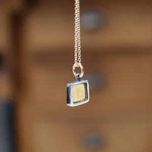 May include: A square pendant necklace featuring a gold-toned center set within a dark, oxidized silver-colored frame. The pendant hangs from a delicate, thin gold-toned chain attached by a small circular jump ring, creating a simple, geometric design.