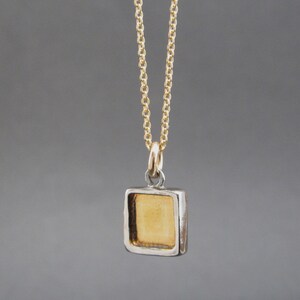 May include: A gold-colored square pendant with a recessed center, suspended from a delicate gold-tone chain. The pendant features a metallic gold interior framed by a silver-toned border, attached to the chain by a small circular jump ring.