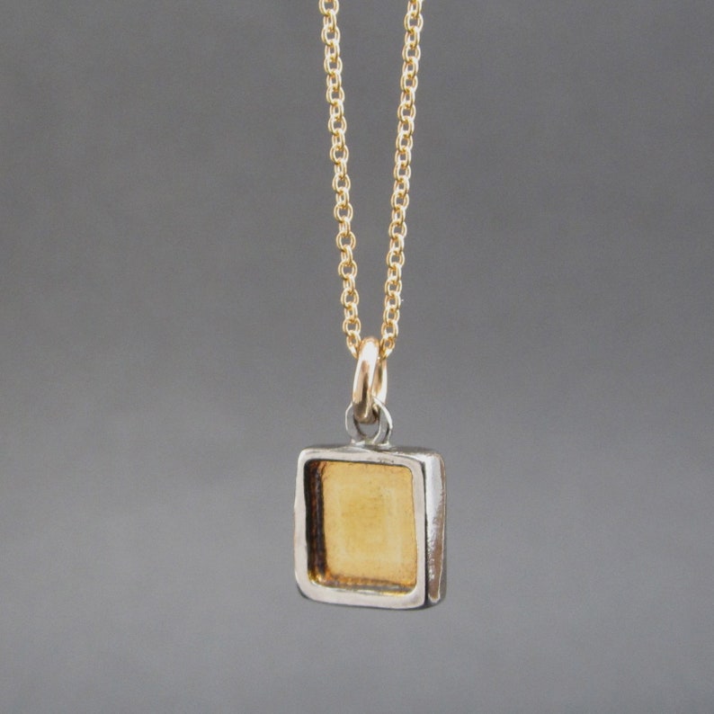 May include: A gold-colored square pendant with a recessed center, suspended from a delicate gold-tone chain. The pendant features a metallic gold interior framed by a silver-toned border, attached to the chain by a small circular jump ring.