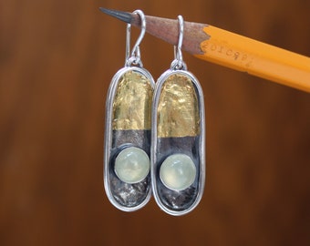 May include: A pair of silver earrings with a unique design. Each earring features a gold-colored top section, a dark gray middle section, and a light green, round stone at the bottom. The earrings are suspended from silver hooks.
