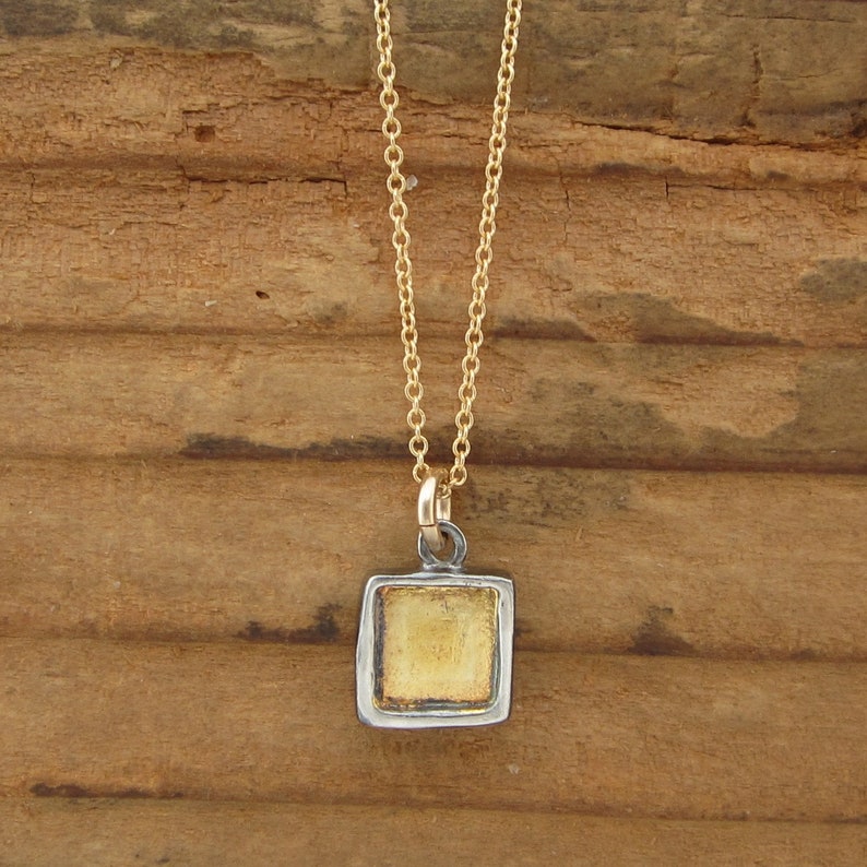 May include: A gold chain necklace featuring a square pendant with a recessed center. The pendant has a dark, oxidized silver-toned border surrounding a textured, metallic gold-colored square inlay, suspended from a delicate gold-toned metal chain.