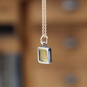 May include: A gold-colored rectangular pendant with a dark metallic border hangs from a delicate gold chain. The pendant features a recessed center with a textured, shimmering gold surface, suspended by a small circular jump ring connector.