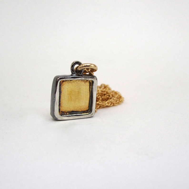 May include: A square pendant necklace with a gold-colored center and a silver-colored frame. The pendant is attached to a delicate gold chain. The necklace is displayed on a white background.