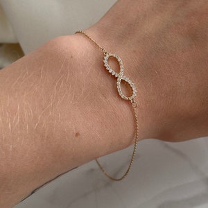 Puede incluir: Un brazalete de oro con un símbolo de infinito con pequeñas piedras transparentes engastadas en el metal. El brazalete está en la muñeca de una persona.