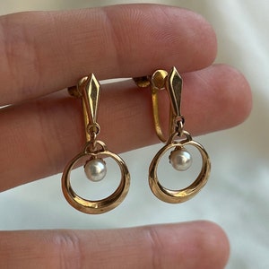 May include: A pair of gold-tone clip-on earrings with a pearl accent. The earrings feature a circular design with a small pearl in the center.