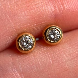 May include: A pair of gold stud earrings, each featuring a round, clear gemstone set in a bezel. The earrings have a simple, classic design with a polished gold finish, suitable for everyday wear.