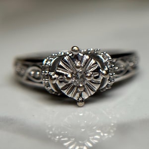 May include: A silver ring with a large, round, clear diamond set in a detailed, ornate design. The ring is set on a white surface.