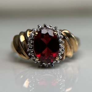 May include: A gold ring featuring a large, oval-shaped, red gemstone surrounded by smaller, white gemstones.
