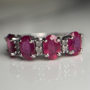 May include: A silver ring with five oval, vivid red gemstones, possibly rubies, and small, clear accent stones. The ring is set on a reflective surface, enhancing the gemstones' colour and the ring's design. The ring is a piece of jewellery.