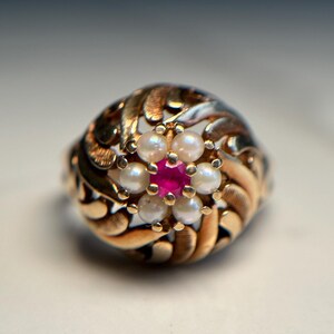 May include: A gold ring featuring a red gemstone surrounded by white pearls. The ring has an intricate swirling design.