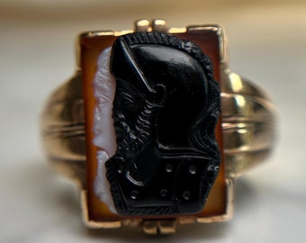 May include: A gold ring with a black and white cameo featuring a profile of a person wearing a helmet. The cameo is set in a rectangular frame.