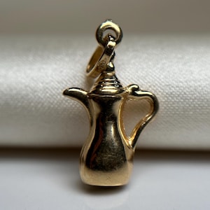 Peut inclure: Un charm doré en forme de cafetière traditionnelle. Le charm possède une anse, un bec verseur et un dessus décoratif. Il est suspendu à une petite boucle, prêt à être ajouté à un collier ou un bracelet. Le charm est probablement en or.